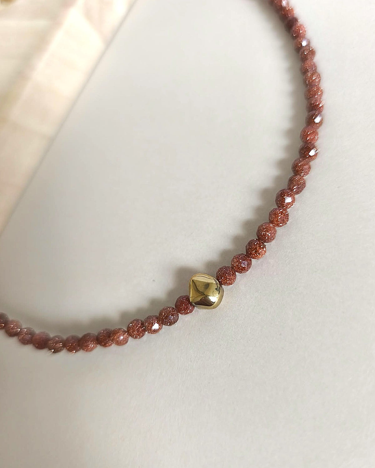A close-up of a beaded necklace with faceted reddish-brown beads and a small polished gold heart-shaped charm, resting on a light neutral surface.